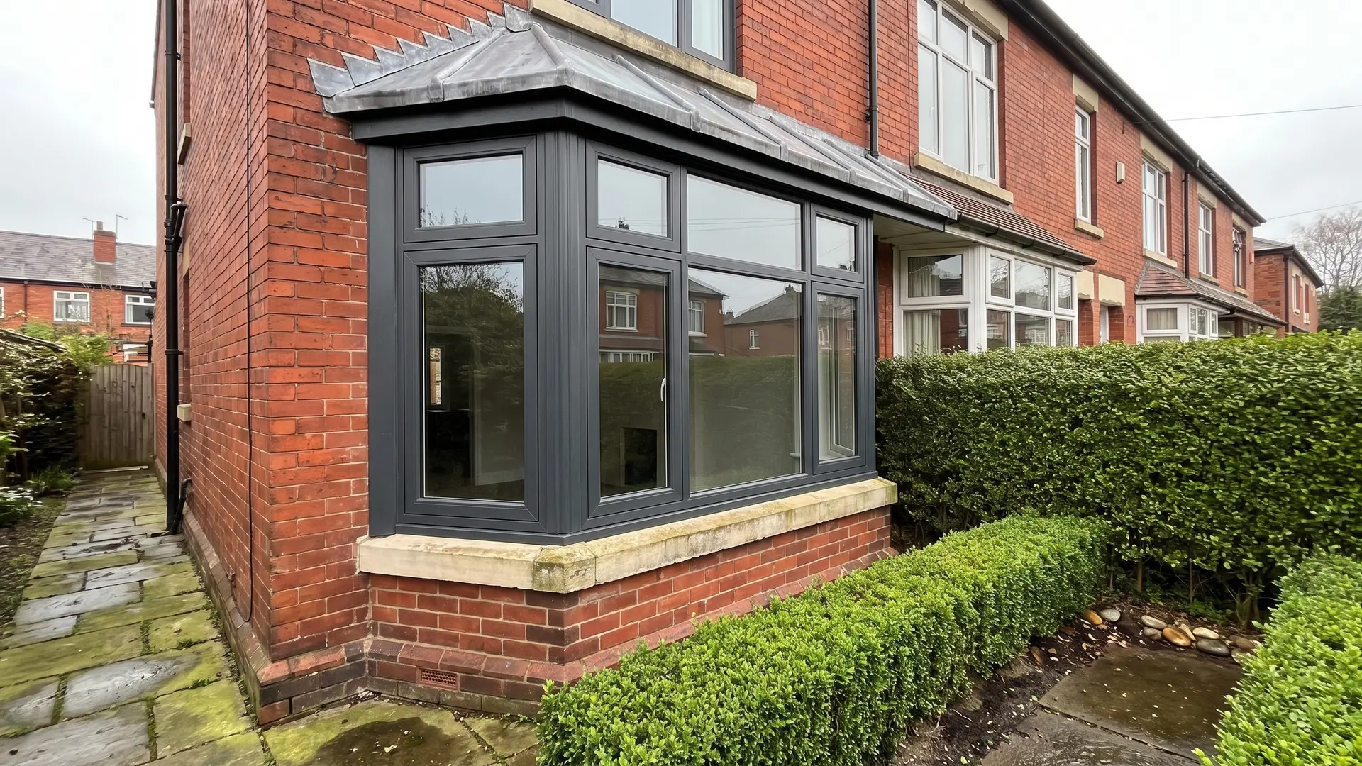 Anthracite grey uPVC casement bay window with angled side returns, a lead-covered bay roof and a stone sill on a red brick terraced house