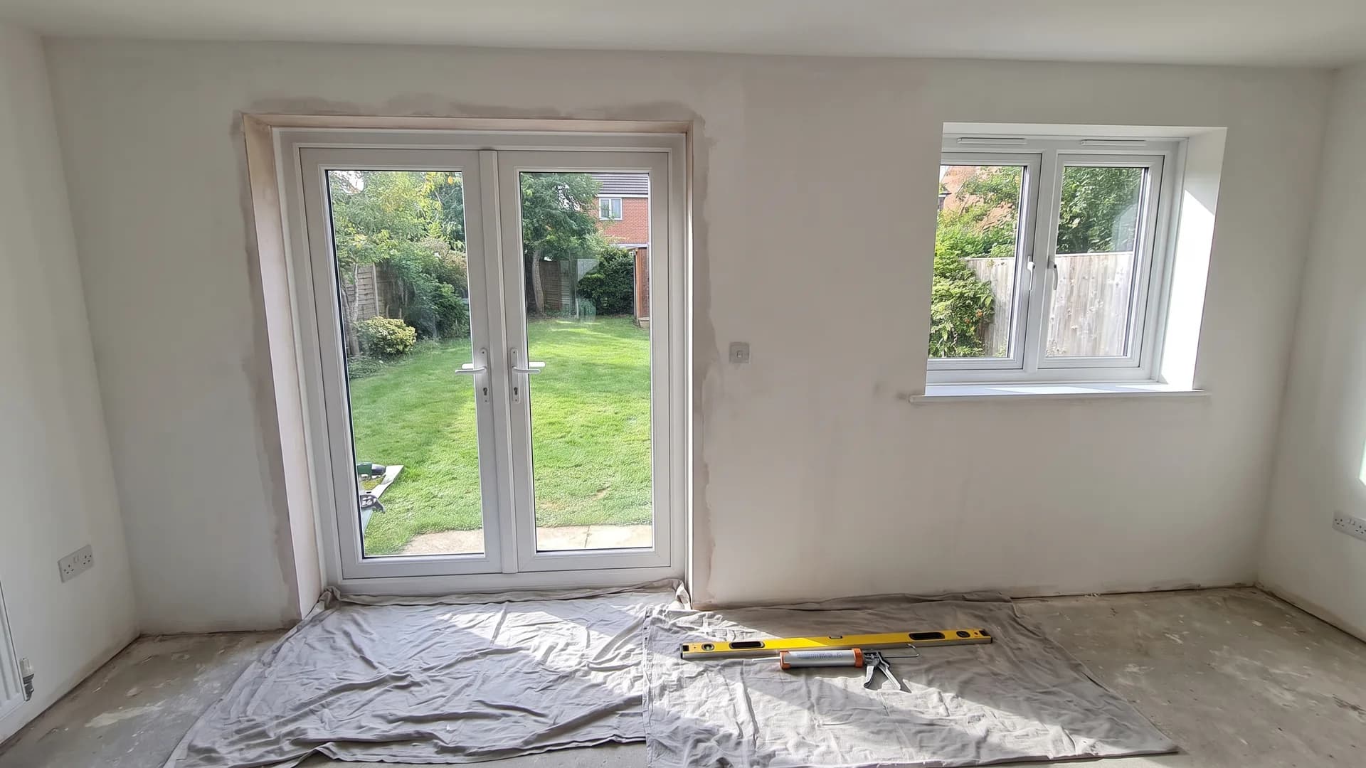 Newly fitted white uPVC French doors and a casement window in a kitchen-diner, with dust sheets, a spirit level and a sealant gun on the floor
