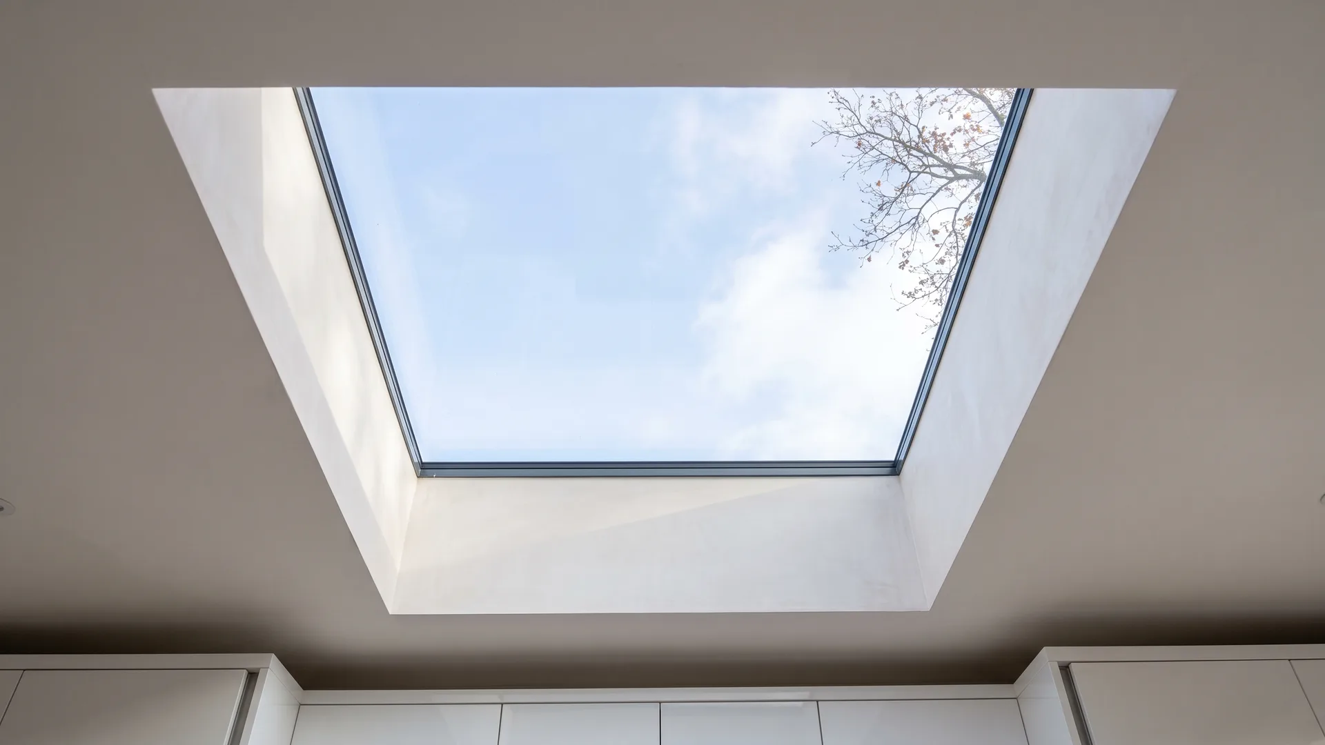 View looking up through a single-panel flat rooflight with a slim anthracite frame and a deep white plastered reveal in a kitchen extension ceiling, pale blue sky and a bare branch beyond