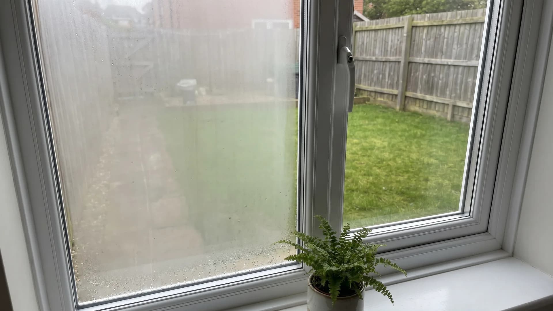 White uPVC casement window seen from inside a living room, with fog trapped between the panes of the left sealed unit and a clear view of the garden through the right pane