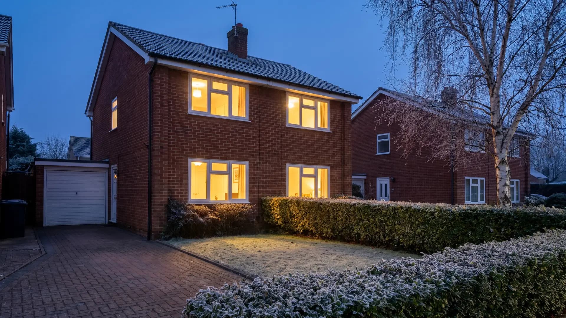 Red brick house at dusk on a frosty evening, its white uPVC double glazed windows lit warmly from inside, with frost on the front lawn and hedge