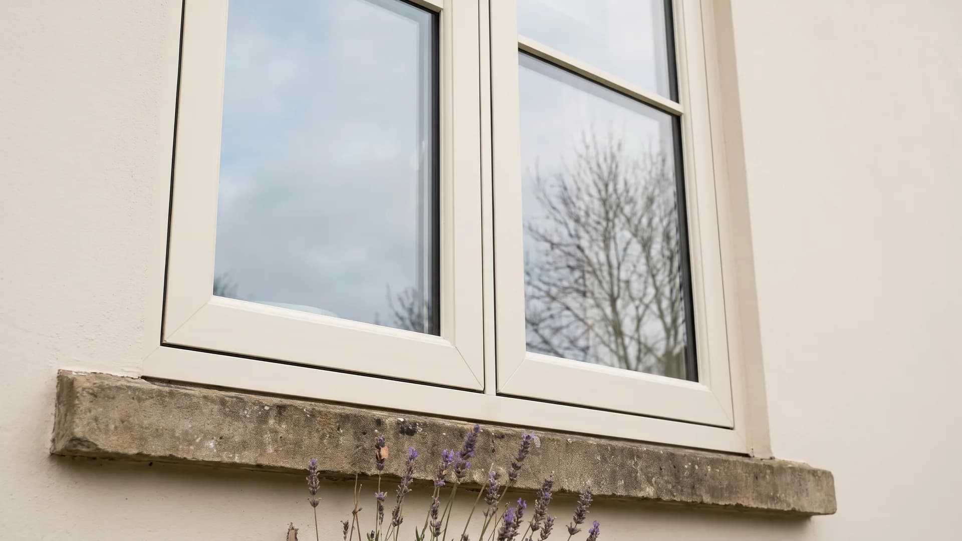 Lower half of a closed cream uPVC flush casement window with flat-faced frames above a weathered stone sill on a pale rendered wall, with lavender in front